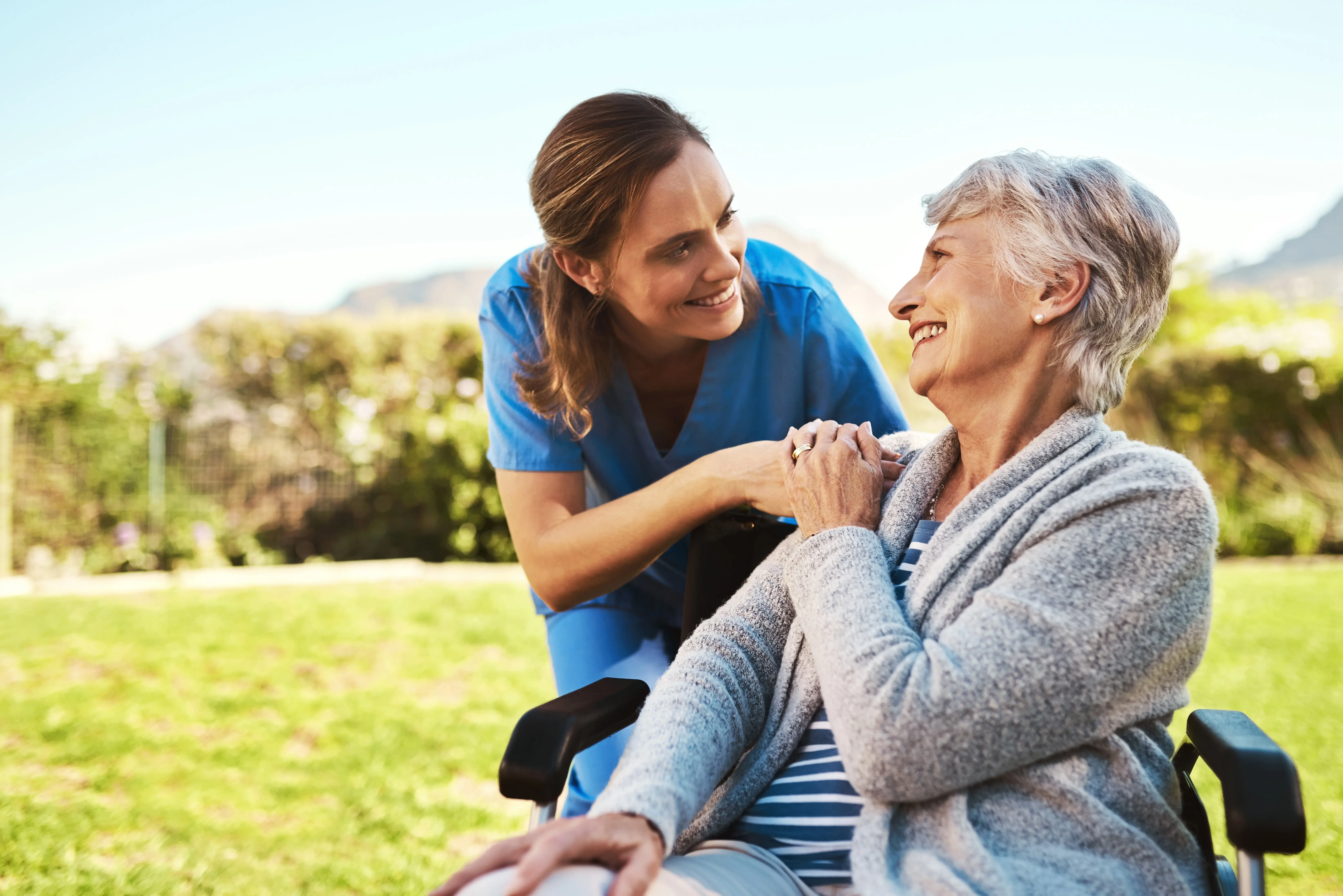 A woman in scrubs smiling at an elderly woman in a wheelchair while holding her hand.